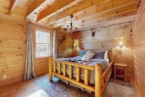 A wooden cabin bedroom with a bed, a window, a chandelier, and a small table. - Charming Log Cabin-  Boat, fish, hike & snowmobile—minutes to lakes & trails (Friendship)