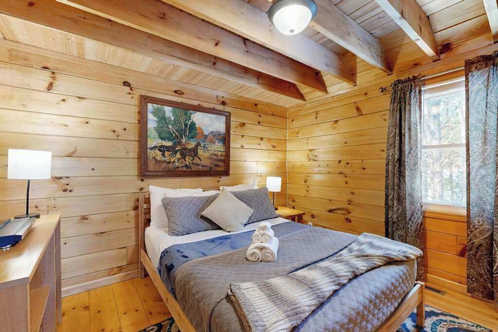 A wooden cabin bedroom with a bed, two bedside tables, a painting on the wall, and a window with curtains.
