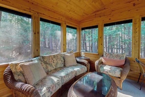 Interior de una cabaña de madera con sofá, una mesa redonda y grandes ventanales que dan a un bosque.