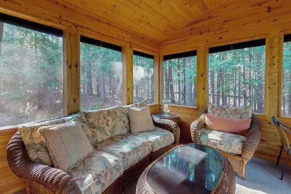 A wooden cabin interior with a sofa, a round table, and large windows offering a forest view.