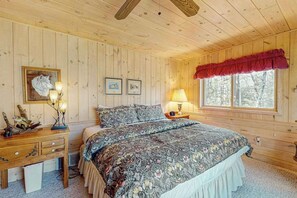 Ein Schlafzimmer einer Holzhütte mit einem Bett, einem Nachttisch, einer Kommode und einem Fenster mit roter Gardine.