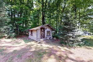 A small log cabin with a wooden door and window, surrounded by dense forest. - Lakefront home with large patio, docks, gas grill, fire pit, & WIFI (Phelps)