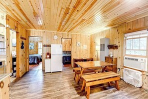 A wooden cabin interior with a dining table, benches, a refrigerator, a fan, and a window.