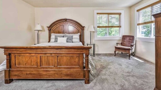 A bedroom with a wooden bed, two bedside lamps, a chair, a window with blinds, and a carpeted floor. - Large lakefront lodge w/Kayaks, Arcade games, WIFI, next to public boat launch (Rhinelander)