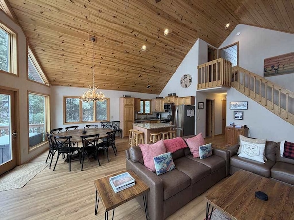 A spacious living area with a high ceiling, wooden flooring, a dining table with chairs, a sofa, and a staircase leading to an upper level.