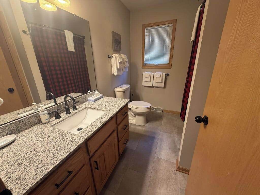 Une salle de bain avec un lavabo sur un plan de travail en granit, des toilettes, une fenêtre avec des stores et un miroir.