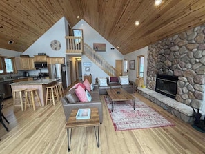 A spacious living area with a stone fireplace, wooden ceiling, and a kitchen with bar stools.