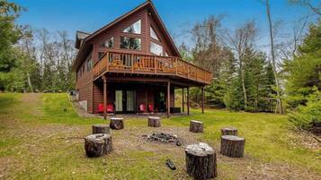 Une cabane en rondins à deux étages, dotée d’un balcon et entourée d’une pelouse où l’on trouve des bûches de bois.