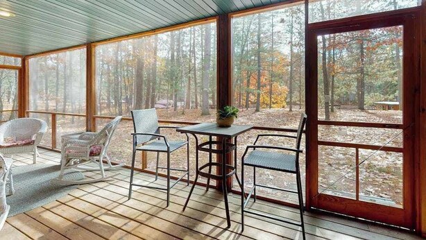 A covered porch with wooden flooring, metal chairs, a table with a plant, and large windows offering a view of a forest. - Riverfront home on the Manitowish Riverway w/ fire pit, kayaks & boat house (Manitowish Waters)