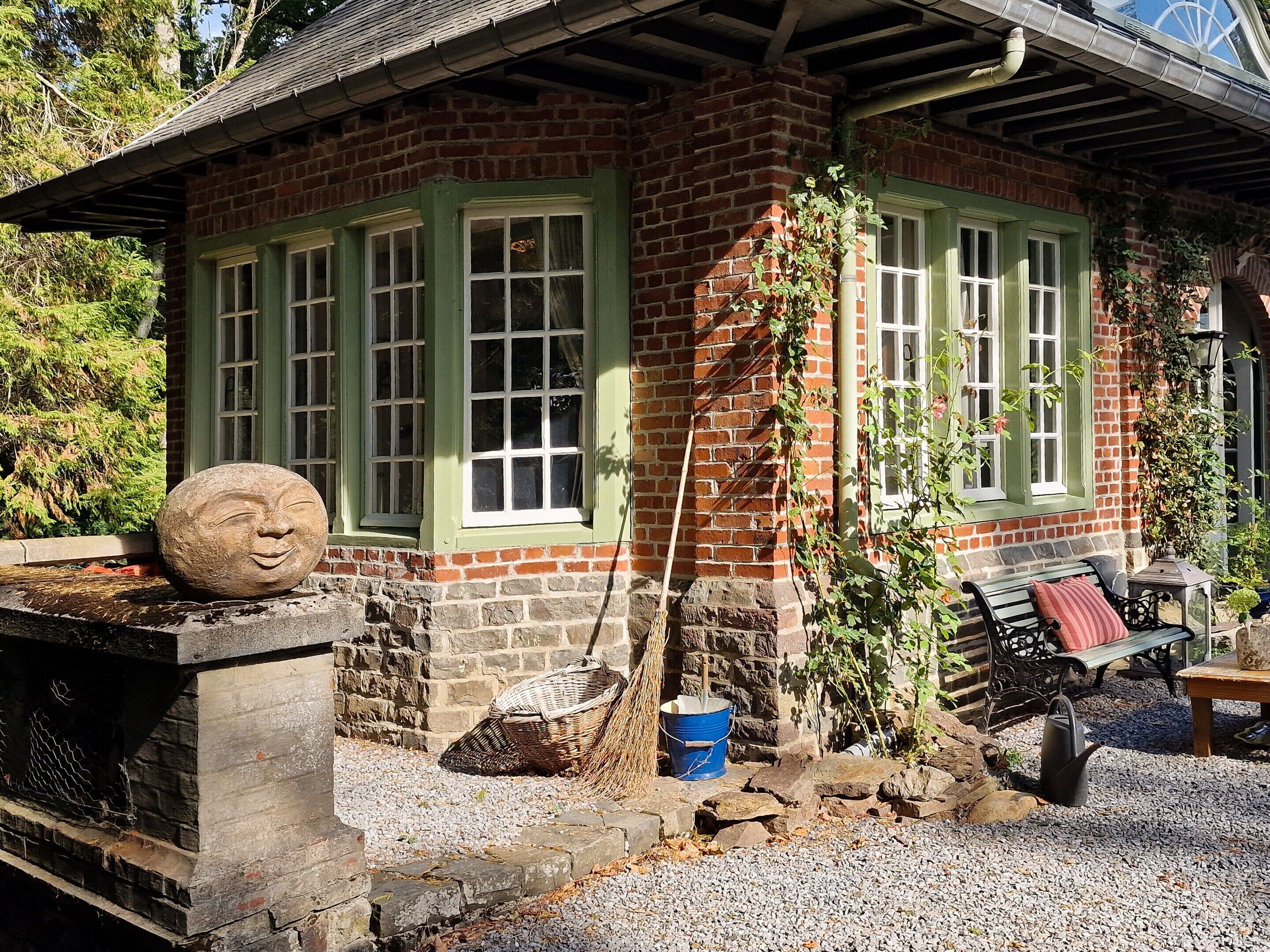 Une maison en briques avec des encadrements de fenêtres verts, une sculpture en pierre sur la façade, un balai, un seau bleu et un banc.