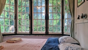 Een bed met een sprei en kussens, een raam met gordijnen, en uitzicht op groen buiten.