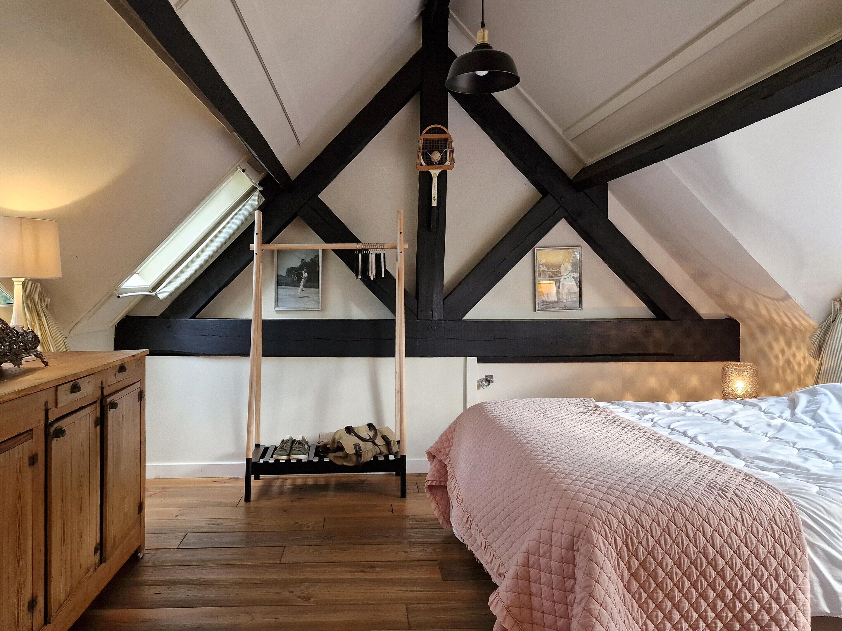 Quarto com teto inclinado, vigas de madeira, cama com colcha rosa, cômoda de madeira e um cabideiro com roupas.