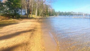 Une plage de sable fin bordée d’une étendue d’eau claire, avec des arbres et un bâtiment visible au loin.