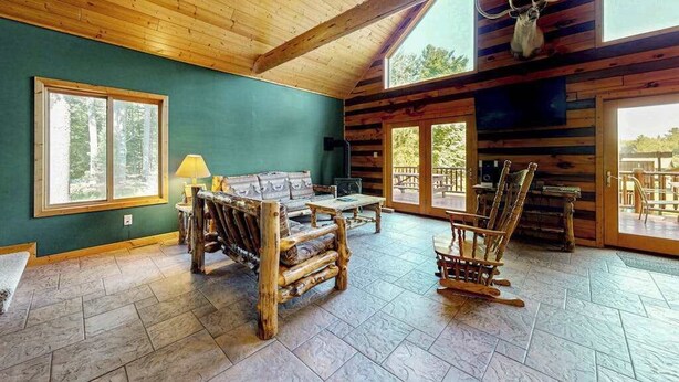 Interior de cabana de madeira com teto de madeira, uma janela grande, um sofá, uma mesa de centro e cabeça de animal montada.