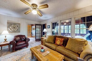 A living room with a ceiling fan, a brown leather armchair, a beige sofa, and a wooden coffee table. - Riverside Home with Firepit,  Gas Grill,  and Washer/Dryer (Manitowish Waters)