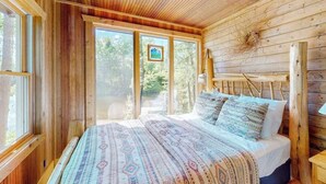 Interior de una cabaña de madera con cama, ventanas amplias y una lámpara en el techo.