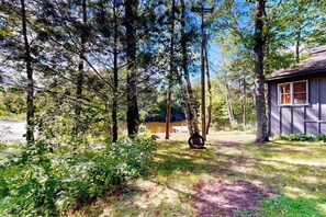 A cabin with a window, surrounded by tall trees and a path leading to it. - Riverside Home with Firepit,  Gas Grill,  and Washer/Dryer (Manitowish Waters)