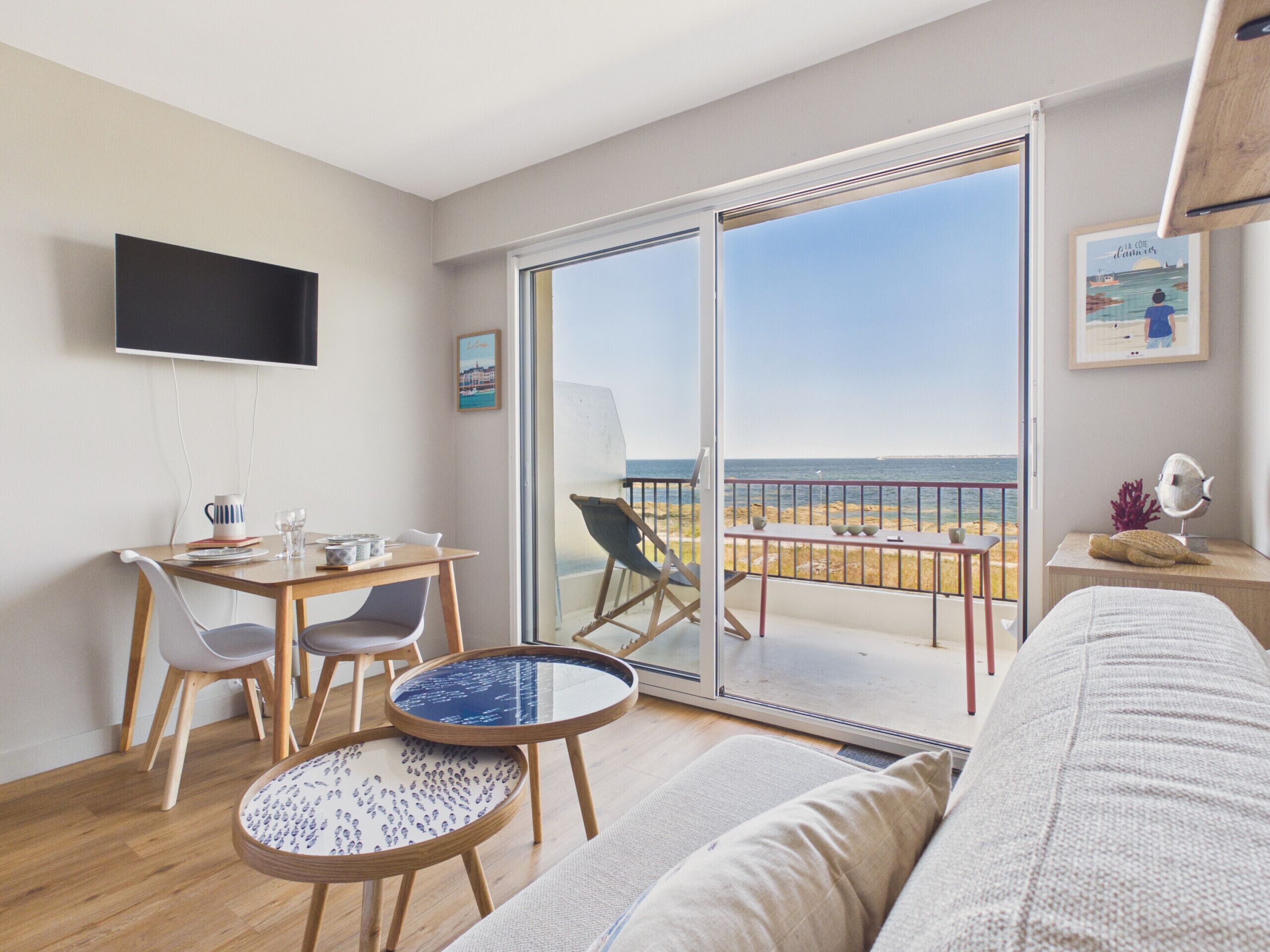 A modern living room with a balcony overlooking the sea, a flat-screen TV mounted on the wall, and a dining area with a table and chairs.