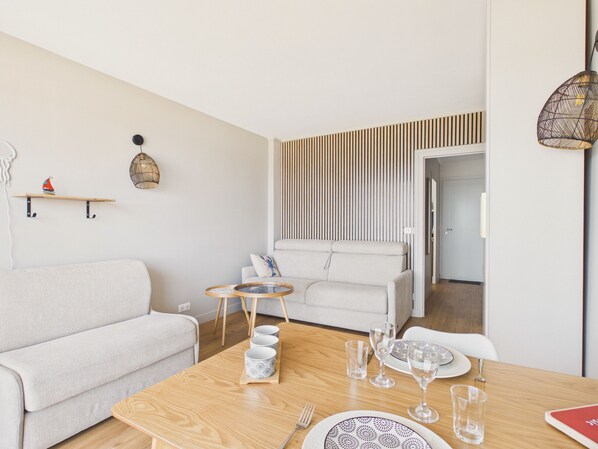 A modern living room with a sofa, a dining table set for four, and a decorative wall panel. - Apartment with sea view 100m from the beach (Le Croisic)