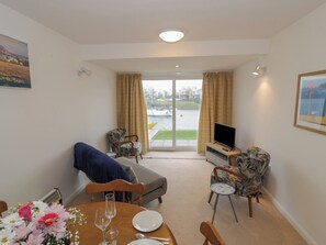 A living area with a sofa, two armchairs, a dining table with flowers, a TV, and a view of a body of water through a glass door. - Harbourside (Porthmadog)