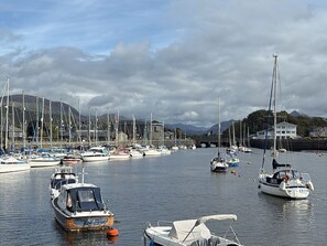 Una marina con varias embarcaciones amarradas, un puente al fondo y un paisaje montañoso.