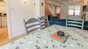 A dining area with a table covered by a tablecloth with animal prints, two chairs with decorative backs, a staircase in the background, and a kitchen visible through an open door. - September Cottage (Weymouth)