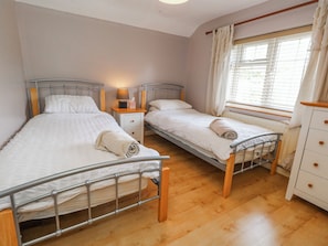 A bedroom with two metal-framed beds, wooden headboards, a bedside table with a lamp, and a window with blinds. - September Cottage (Weymouth)