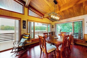 A dining area with a wooden table and chairs, large windows, and a view of greenery.