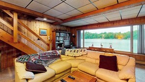 Interior acogedor de una cabaña con escalera de madera, sofá modular, estantería y un gran ventanal con vistas a un lago.