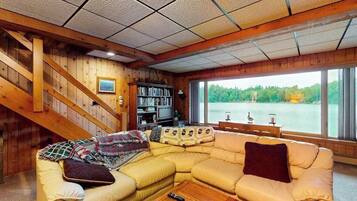 L’intérieur d’un chalet accueillant, avec un escalier en bois, un canapé d’angle, une bibliothèque et une grande fenêtre donnant sur un lac.