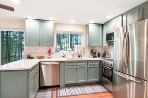 A modern kitchen with grey cabinets, stainless steel appliances, and a large window overlooking a forest. - Lakefront, Dog-Friendly Cabin with Lake View, Central AC, Dock, Deck, Free WiFi (Minocqua)
