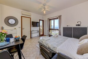 A bedroom with a bed, a desk with a chair, a dining table with chairs, a TV, and a view of the sea. - MGH - My Lakeview Escape (Peschiera del Garda)