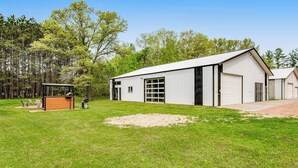 A single-story building with a large garage door, a covered outdoor seating area, and a grassy yard. - Barn & cabin with firepits, grills, foosball, kayaks, home theater, & 2 kitchens (Necedah)