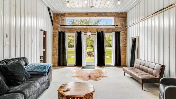 A spacious room with a large window, a leather sofa, a wooden coffee table, and a stone fireplace. - Barn & cabin with firepits, grills, foosball, kayaks, home theater, & 2 kitchens (Necedah)