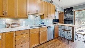 A modern kitchen with wooden cabinets, a stainless steel dishwasher, and a bar area with stools. - Green side smart home with golf, beach/lake access, off-site dock & EV charger (Nekoosa)