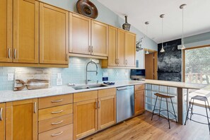 A modern kitchen with wooden cabinets, a stainless steel dishwasher, and a bar area with stools. - Green side smart home with golf, beach/lake access, off-site dock & EV charger (Nekoosa)