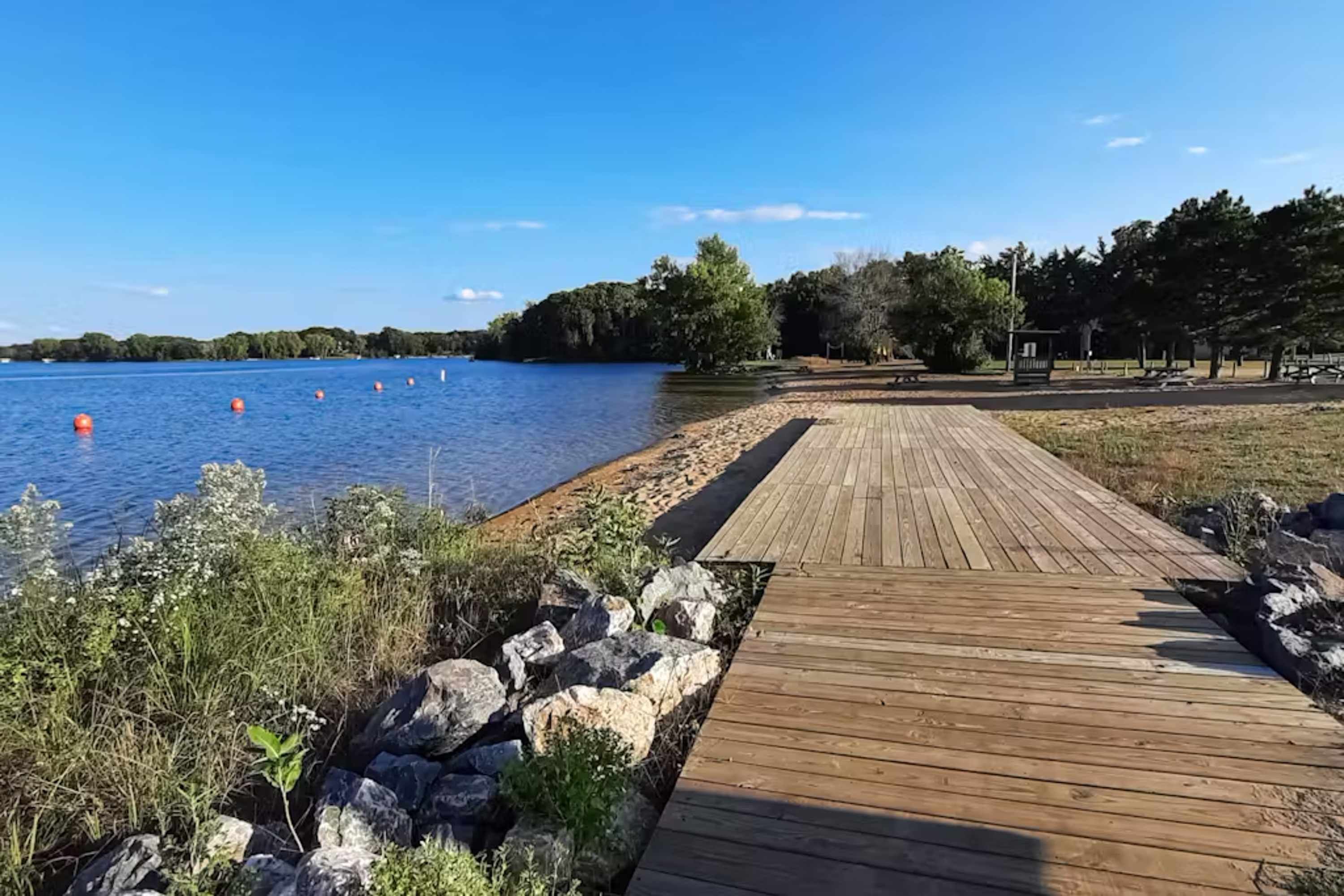 Une promenade en bois menant à un lac où flottent des bouées orange, le tout étant entouré de rochers et de végétation.