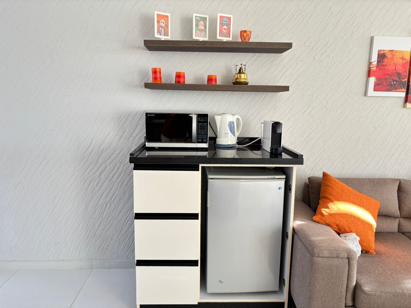 Une petite cuisine équipée avec un micro-ondes, une bouilloire et une machine à café sur un plan de travail noir, un mini-réfrigérateur et un canapé agrémenté d’un coussin orange.