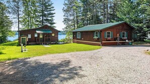 A cabin with a gravel driveway, a wooden deck, and a view of a lake surrounded by trees. - Cozy, lakefront cottage w/ a firepit,kayak,shared patio, gas grill, & free WiFi (Eagle River)