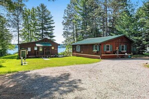 A cabin with a gravel driveway, a wooden deck, and a view of a lake surrounded by trees. - Cozy, lakefront cottage w/ a firepit,kayak,shared patio, gas grill, & free WiFi (Eagle River)