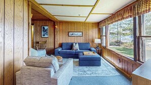 A cozy living room with wooden walls, a blue sofa, and a large window with a view of trees. - Cozy, lakefront cottage w/ a firepit,kayak,shared patio, gas grill, & free WiFi (Eagle River)
