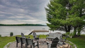 A lakeside seating area with Adirondack chairs, a fire pit, and a dock extending into the water. - Lakefront home with private dock, firepit, playground & lake views (Eagle River)