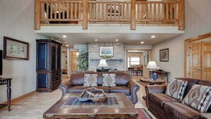 A spacious living room with a high ceiling, a stone fireplace, and a wooden balcony. - Lakefront home with private dock, firepit, playground & lake views (Eagle River)