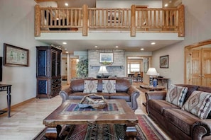 A spacious living room with a high ceiling, a stone fireplace, and a wooden balcony. - Lakefront home with private dock, firepit, playground & lake views (Eagle River)
