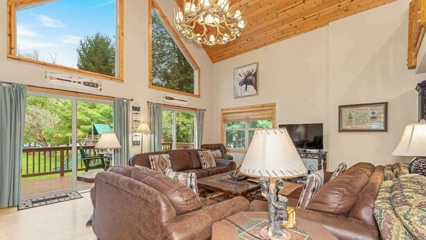 A spacious living room with leather sofas, a chandelier, and large windows offering a view of the outdoors. - Lakefront home with private dock, firepit, playground & lake views (Eagle River)