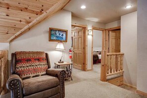 A cozy room with a sloped ceiling, a patterned armchair, a small table with a lamp, and wooden doors leading to other areas.