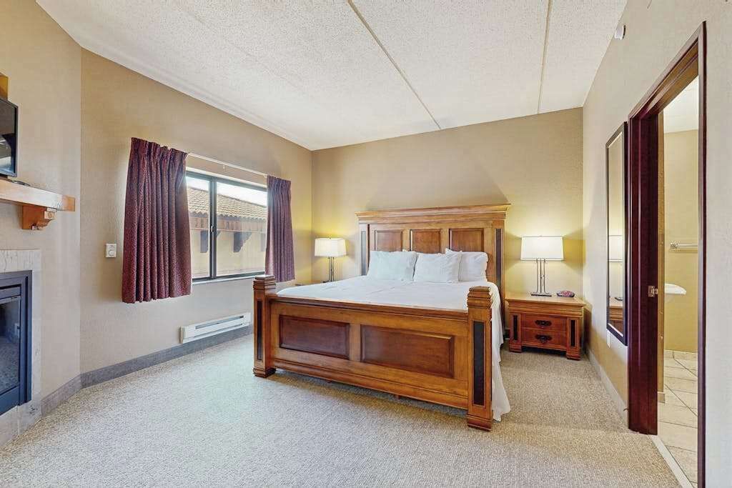 A bedroom with a wooden bed, a nightstand, a lamp, a window with curtains, and a door.