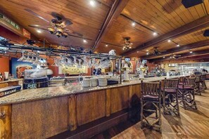 Zona de bar con techo de madera, taburetes y una barra con vasos y botellas.