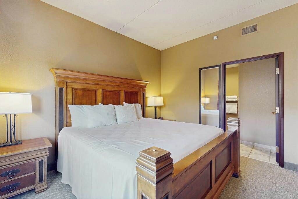 A hotel room with a large bed, bedside lamps, a wooden headboard, a wardrobe, and a door to another room.