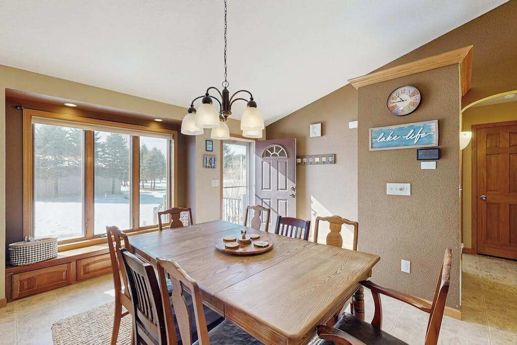 A dining area with a wooden table, chairs, a chandelier, and a view through large windows.